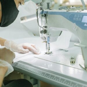 A woman wearing a face mask and gloves operates a sewing machine in a workshop.